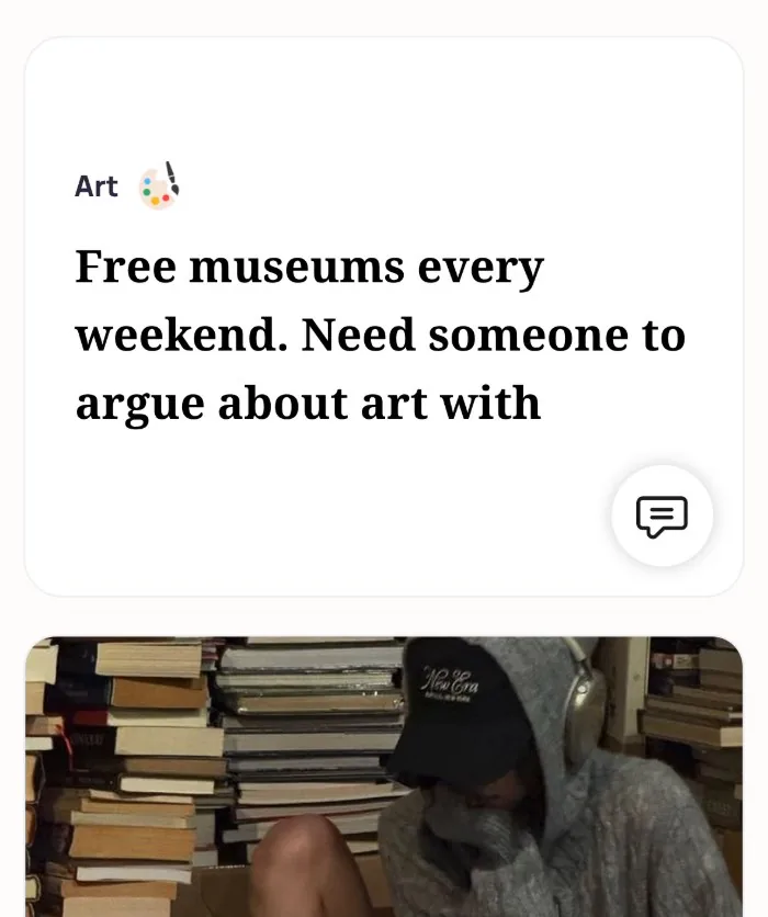 An Alyke profile card for Art that reads: Free museums every weekend. Need someone to argue about art with, above a photo of someone in a hoodie and cap sitting among stacks of books