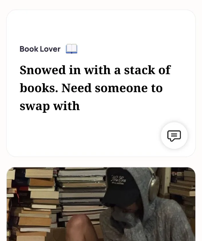 An Alyke profile card for Book Lover that reads: Snowed in with a stack of books. Need someone to swap with, above a photo of someone in a hoodie and cap sitting among stacks of books