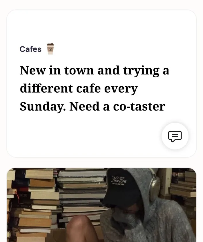 An Alyke profile card for Cafes that reads: New in town and trying a different cafe every Sunday. Need a co-taster, above a photo of the member in a hoodie and headphones among stacks of books