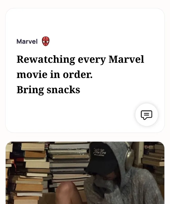An Alyke profile card for Marvel that reads: Rewatching every Marvel movie in order. Bring snacks, above a photo of someone in a hoodie and cap sitting among stacks of books