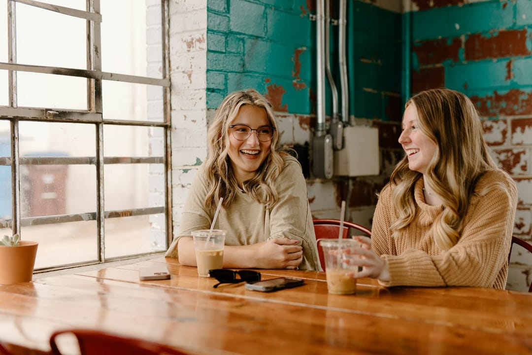 Two new friends meeting at a cafe counter, one waving as she walks in
